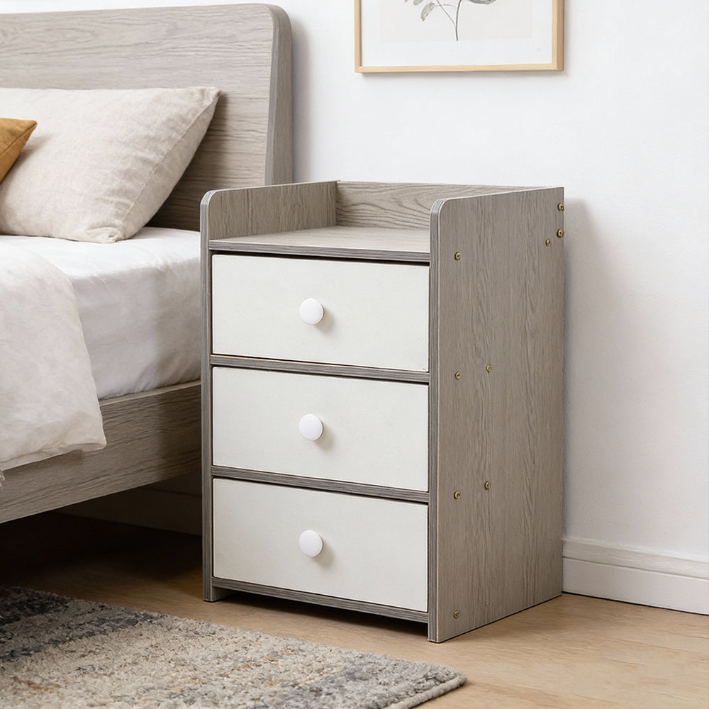 Grey and white bedside table with 3 drawers next to a bed with a plant on top