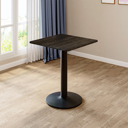 Black square pedestal table with dark wood-grain top and round metal base standing by a window on oak flooring