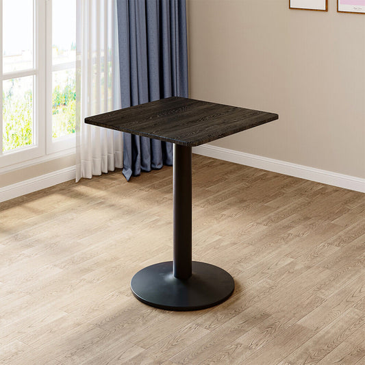 Black square pedestal table with dark wood-grain top and round metal base standing by a window on oak flooring