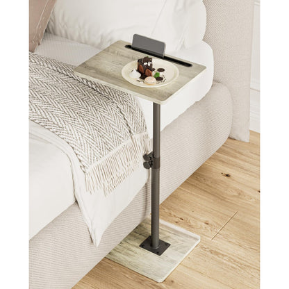 Grey wood effect height adjustable side table next to a bed with a plate of dessert