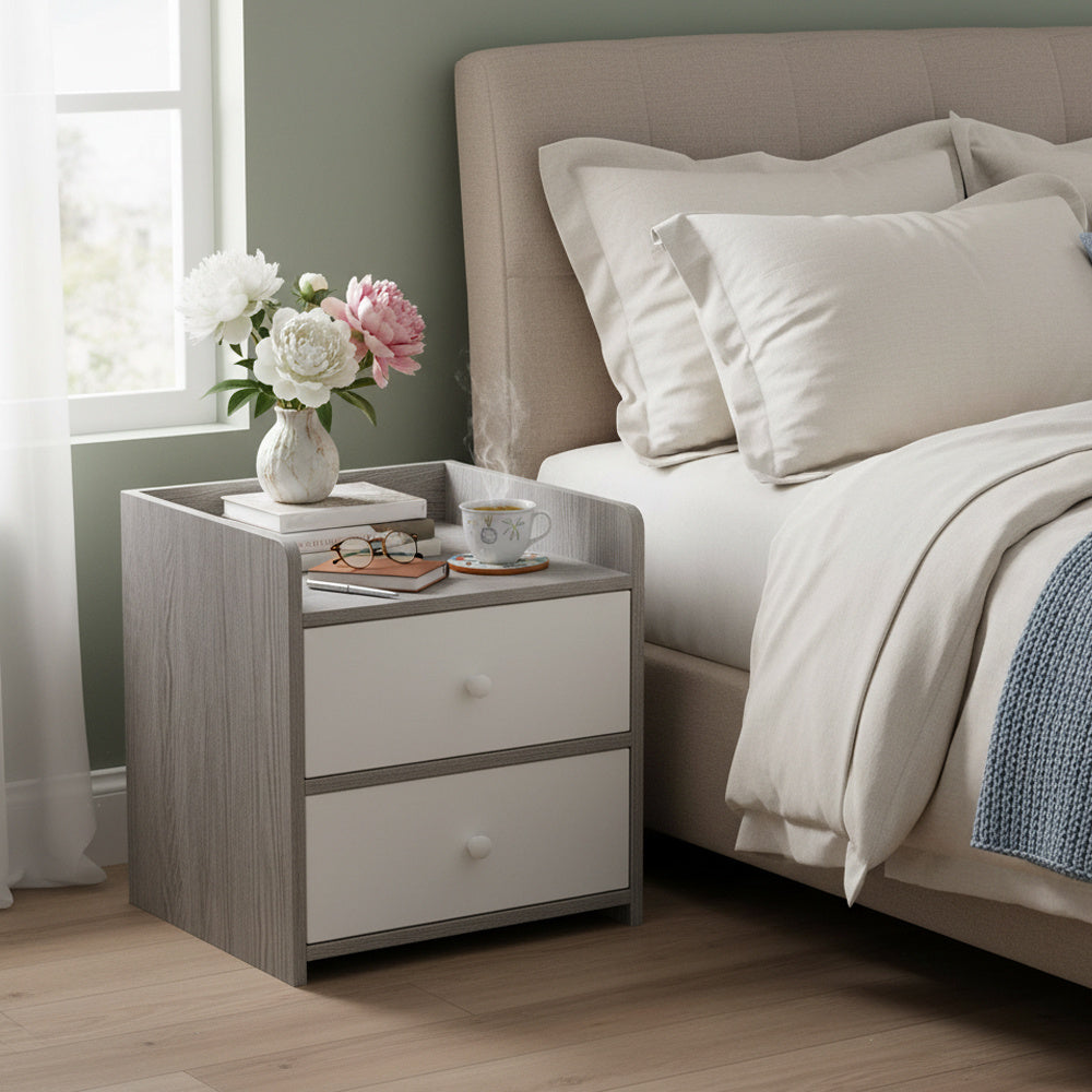 Grey wood-effect nightstand with white drawer beside a bed with flowers and tea