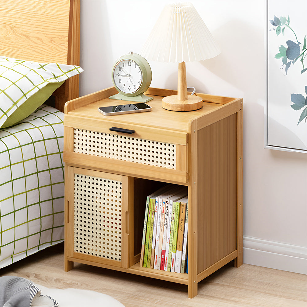 Light wood bedside table with drawer and cupboard beside a bed with a pleated lamp and clock