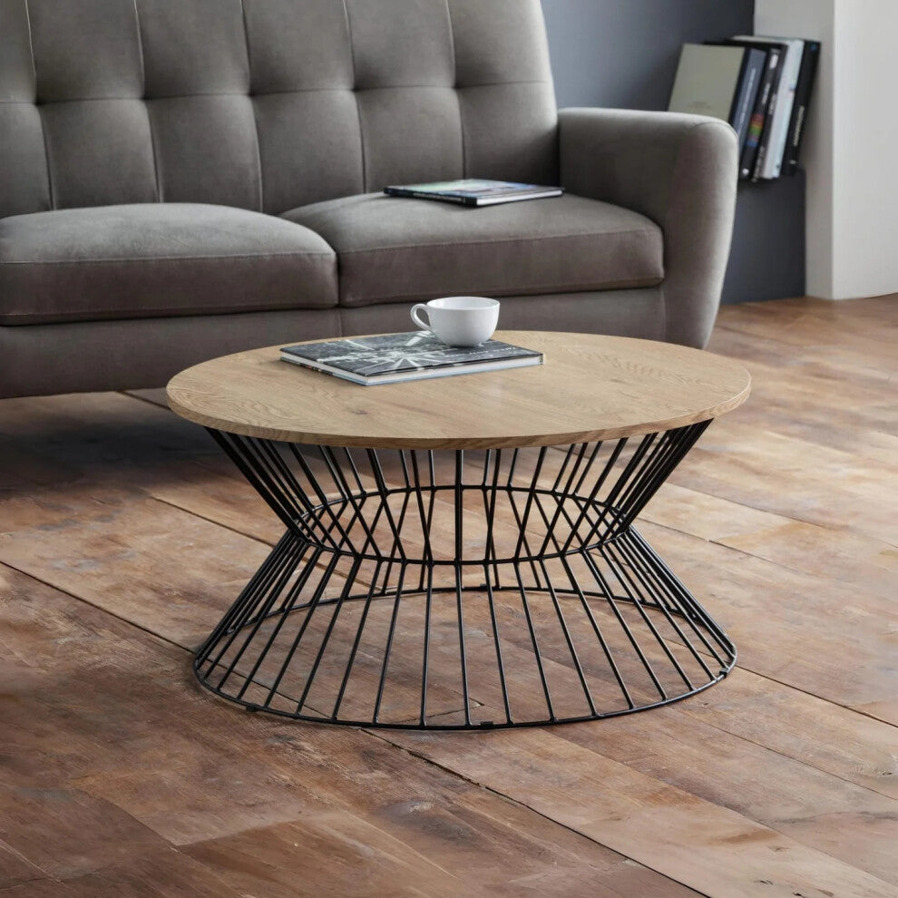 Round oak coffee table with black wire hourglass base in front of a grey sofa on rustic floorboards