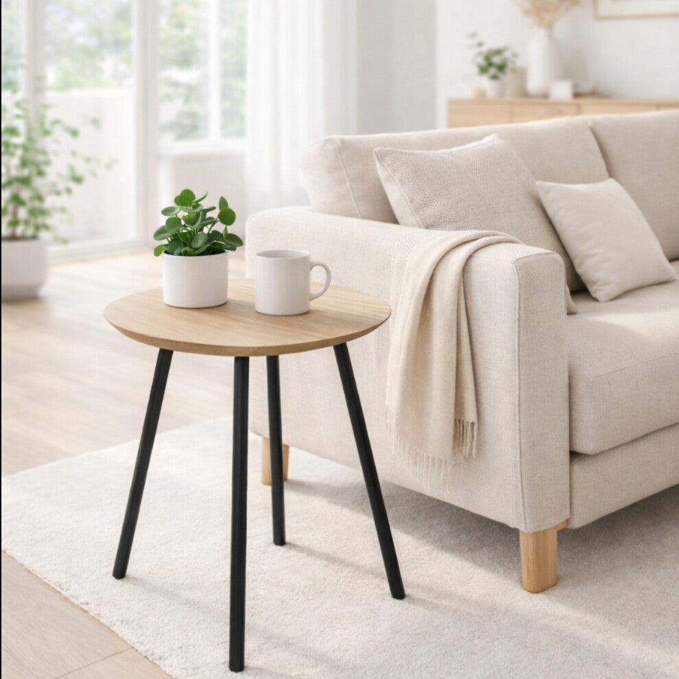 Round side table with black legs and natural wood-effect top holding a plant and mug beside a cream linen sofa