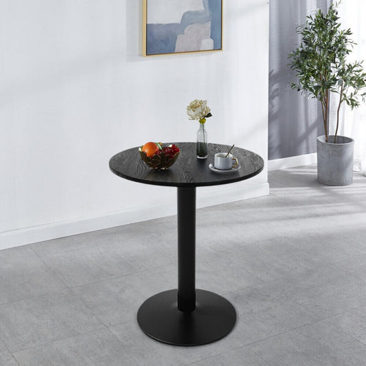 Small round black pedestal table with wood-grain top set with coffee cup, fruit bowl and flower vase in a bright modern room
