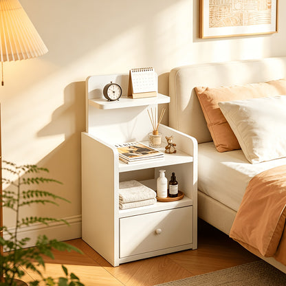 Slim white tiered bedside table with drawer beside a bed with clock and books